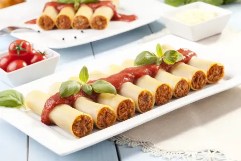 Cannelloni z mięsem i sosem pomidorowym, udekorowane bazylią, na białym talerzu.