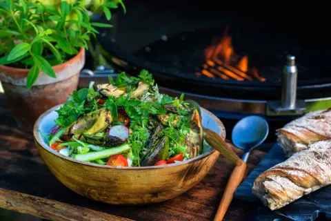 En härlig somrig sallad med grillade grönsaker, tomater och rädisor i en träskål. Bredvid ligger nybakat bröd och en grill med flammor i bakgrunden.