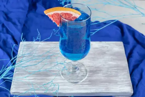 Niebieski drink, jak orzeźwiająca blue lagoon, z plasterkiem grejpfruta, na drewnianej desce, otoczony błękitnymi gałązkami.