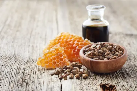 Propolis co to? Miód w plastrze, drewniana miseczka z propolisem i buteleczka z ciemnym płynem na drewnianym tle.