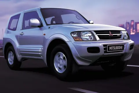 Srebrny Mitsubishi Shogun 3-drzwiowy na drodze o zmierzchu, z miejskim krajobrazem w tle.