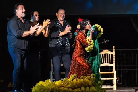 Una bailaora de flamenco andalusi, con un vestido rojo y amarillo, besa un mantón verde. A su lado, tres músicos aplauden.