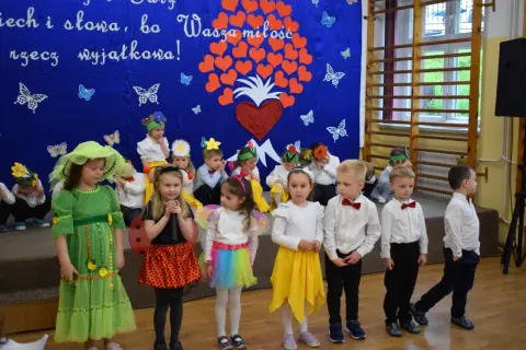 Przedszkolanka w zielonej sukience i dzieci w strojach na tle niebieskiej kurtyny z sercami i motylami.