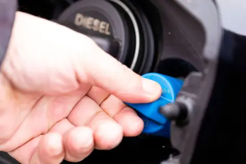 Hand schraubt blauen AdBlue-Deckel ab. Diesel-Tankdeckel daneben.