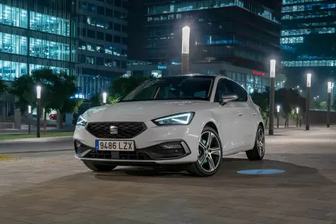 Blanco Seat León aparcado de noche frente a edificios modernos. Las luces LED del coche y las farolas crean un ambiente urbano. Opiniones sobre este modelo son muy positivas.