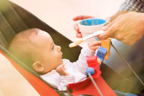 Karmienie 4-miesięcznego niemowlęcia. Zastanawiasz się, ile powinno jeść 4 miesięczne niemowlę? Maluch w leżaczku z zaciekawieniem patrzy na łyżeczkę z jedzeniem.
