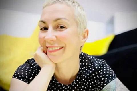 Laura Veltz, a blonde woman with short hair, smiles and rests her chin on her hand. She wears a black polka dot shirt.