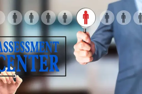 Uomo in giacca blu esamina candidati con lente d'ingrandimento. Testo "ASSESSMENT CENTER" scritto su schermo.
