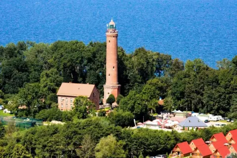 Latarnia morska Sarbinowo góruje nad zielonymi drzewami, z błękitnym morzem w tle. Obok niej budynki i czerwone dachy domków letniskowych.