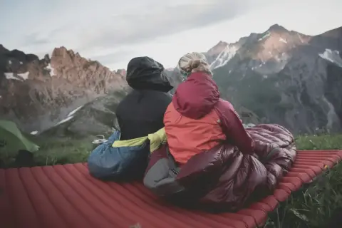 Dos personas sentadas en un colchón inflable, envueltas en sacos de dormir, disfrutan de un paisaje montañoso. El material vivac les permite acampar cómodamente.