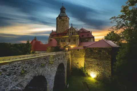Zamek Czocha, gdzie jest, w blasku zachodzącego słońca i nocnych świateł, z kamiennym mostem i wieżą.