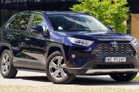Elegancka, granatowa Toyota RAV4 5 generacji z przyciemnianymi szybami i chromowanymi felgami, zaparkowana na tle zieleni i nowoczesnego budynku.