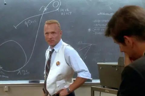 Gene Kranz, w białej kamizelce, omawia trajektorię misji Apollo 13 na tablicy.