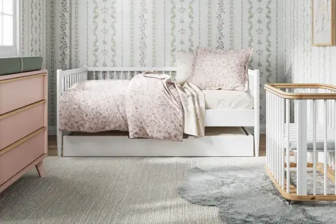 A charming small nursery features a white daybed with floral bedding, a pink dresser, and a white crib.