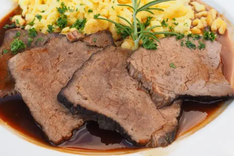 Fertig eingelegter Sauerbraten zubereiten: Zarte Scheiben Rindfleisch in dunkler Sauce mit Spätzle und Rosmarin garniert.