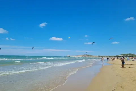 Spiaggia di Scialmarino con kitesurfer che danzano nel cielo azzurro e bagnanti che passeggiano sulla sabbia dorata.