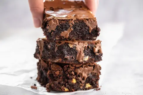 En hand lyfter en stapel saftiga, mörka glutenfria brownies med smält choklad.
