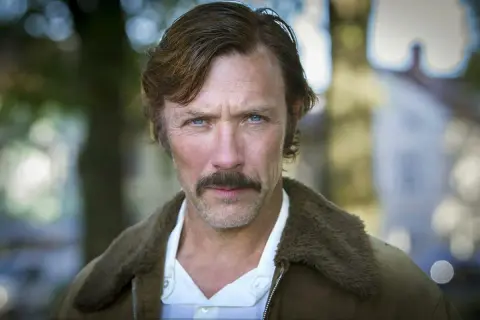 Porträtt av en svensk manlig skådespelare från 2000-talet med mustasch och blå ögon, klädd i en brun jacka med foder.