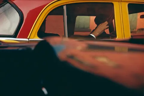 Vue à travers une fenêtre de voiture rouge et jaune, la main d'un homme au volant, typique des saul leiter photos.