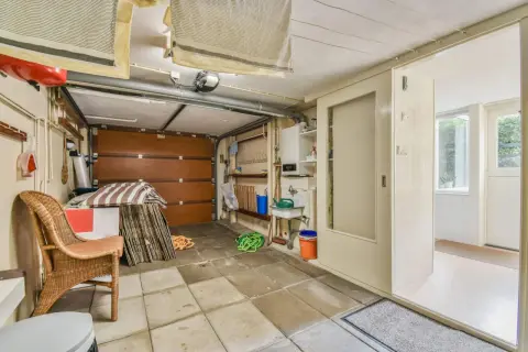 Un garage en cours de transformation en habitation. On y voit une porte de garage, un évier, une chaise en rotin et des éléments de rangement.