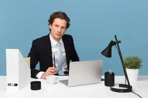 Młody manager w garniturze siedzi przy biurku, trzymając długopis. Na biurku laptop, filiżanka i lampka.