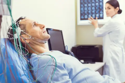 Mężczyzna leży z elektrodami na głowie do badania eeg. Lekarka analizuje wyniki na ekranie.