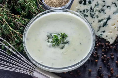 Crema suave y sedosa, lista para tu salsa roquefort receta. Ingredientes frescos como tomillo y pimienta negra completan la escena.