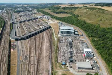 Rozległa infrastruktura kolejowa i stacja transformatorowa przy Eurotunelu. W tle wzgórza z białym koniem.