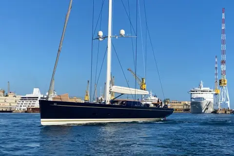 Il kauris iv yacht, scuro e imponente, naviga in un porto con gru e navi da crociera sullo sfondo.