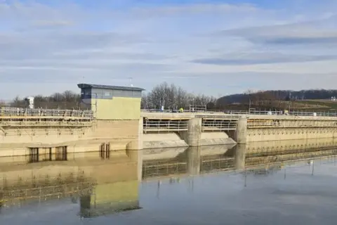 Betonowa zapora w Sieniawie w budowie, z widocznym budynkiem sterowni i rusztowaniami. Woda odbija konstrukcję.
