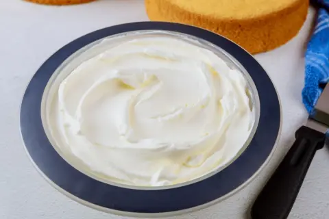 Puszysta bita śmietana z mascarpone w misce, gotowa do dekorowania tortu. Obok leżą biszkopty i szpatułka.