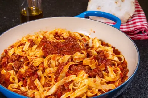 Breite Tagliatelle mit herzhaftem Ragu Bolognese in einer blauen Pfanne. Daneben ein Glas Olivenöl und ein kariertes Tuch.