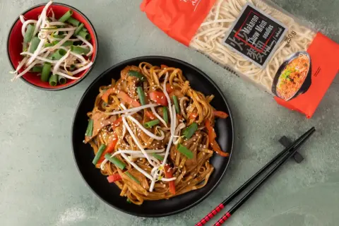Gotowy do przygotowania dania z makaronem chow mein, który jest idealny do ramen. Na talerzu danie z warzywami i mięsem, obok miseczka kiełków i opakowanie makaronu.
