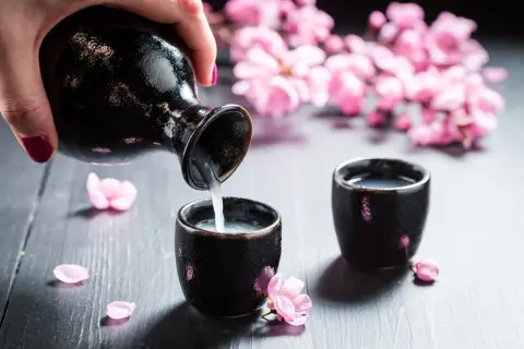 Ręka nalewa sake do czarki. Delikatne płatki wiśni i kwitnące gałązki tworzą wiosenną atmosferę, idealną do nauki, jak pić sake.