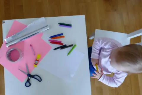 Dziecko przygotowuje materiały do zrobienia lustra z folii aluminiowej: kredki, nożyczki, taśmę, papier i folię.