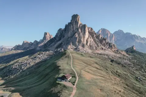 Un refugio alpino solitario en la ladera de una montaña, con un sendero que serpentea hacia él. El paisaje evoca la belleza de **Milano Cortina**, con picos imponentes y cielos despejados.