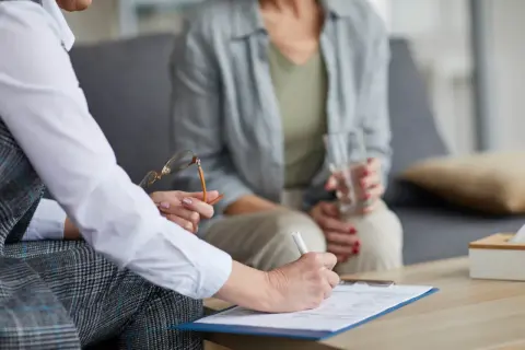 Psychoterapeutka pisze na dokumentach, druga osoba trzyma szklankę wody.