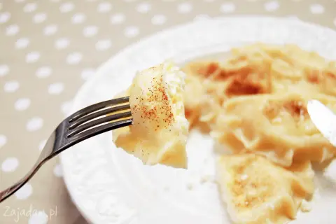 Widok z bliska na widelec z pierogiem z serem, posypanym cynamonem. Na talerzu widać więcej pysznych pierogów.