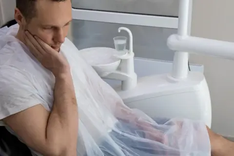 Mężczyzna w fotelu dentystycznym, z ręką na policzku, martwi się o swoje zdrowie. Czy to początek sepsy od zęba?