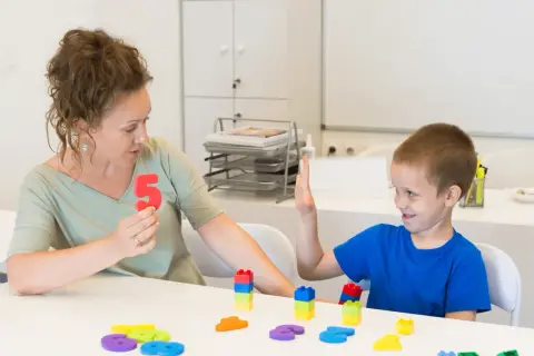 Kobieta pokazuje chłopcu liczbę 5, podczas gdy on podnosi rękę. To scena z zajęć surdopedagogiki, gdzie nauka odbywa się przez zabawę.