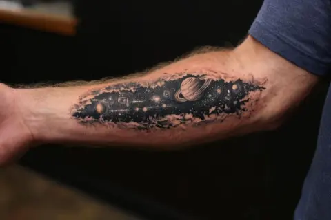 Tatuaż kosmos na przedramieniu: Saturn, planety i gwiazdy w czerni i bieli.