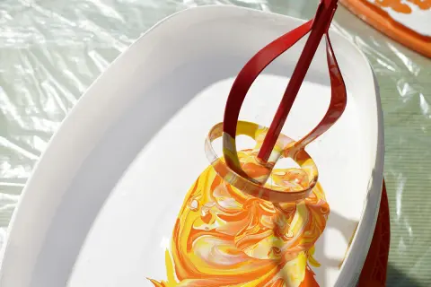 Ein roter Rührer mischt leuchtend gelbe und orangefarbene Wandfarbe in einem weißen Eimer.