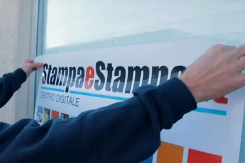 Mani applicano un grande adesivo con logo "Stampa e Stampe Centro Digitale" su una superficie, mostrando come applicare un adesivo senza bolle per un risultato perfetto.