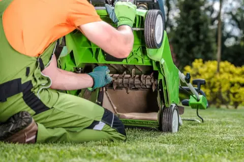 Mężczyzna w zielonym ogrodniczkach i pomarańczowej koszulce sprawdza mechanizm skaryfikatora do trawy.