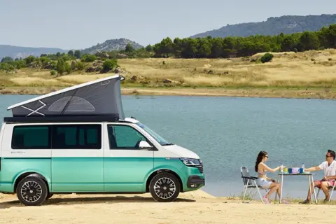 Volkswagen California con baño, lista para una escapada junto al lago. Pareja disfruta de un picnic al aire libre.
