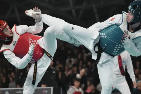 Två taekwondo-utövare i strid. Den ena med röd väst och hjälm, den andra med blå väst och hjälm. Båda utför sparkar.
