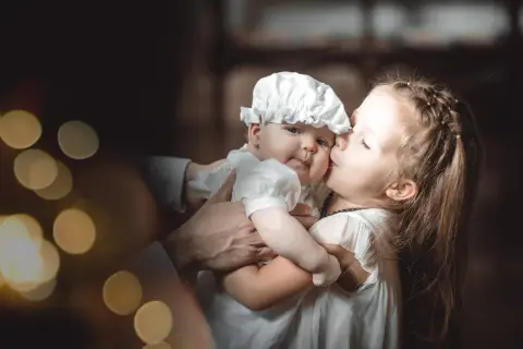 Une sœur embrasse tendrement son bébé, habillé pour baptiser son enfant. Lumières douces et ambiance chaleureuse.