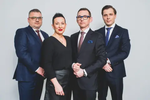 Cztery osoby w garniturach i czarnej bluzce, stojące obok siebie na jasnym tle.