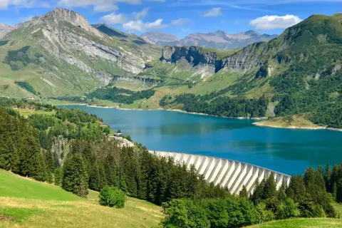 Vue panoramique du lac de Roselend, un barrage imposant bordé de forêts verdoyantes et de montagnes majestueuses sous un ciel bleu éclatant.