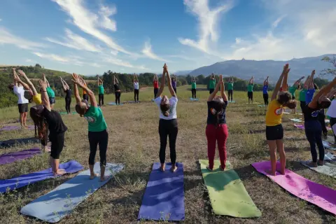 Gruppo di persone in posa urdhva hastasana su tappetini da yoga in un prato verde, con cielo azzurro e nuvole bianche.